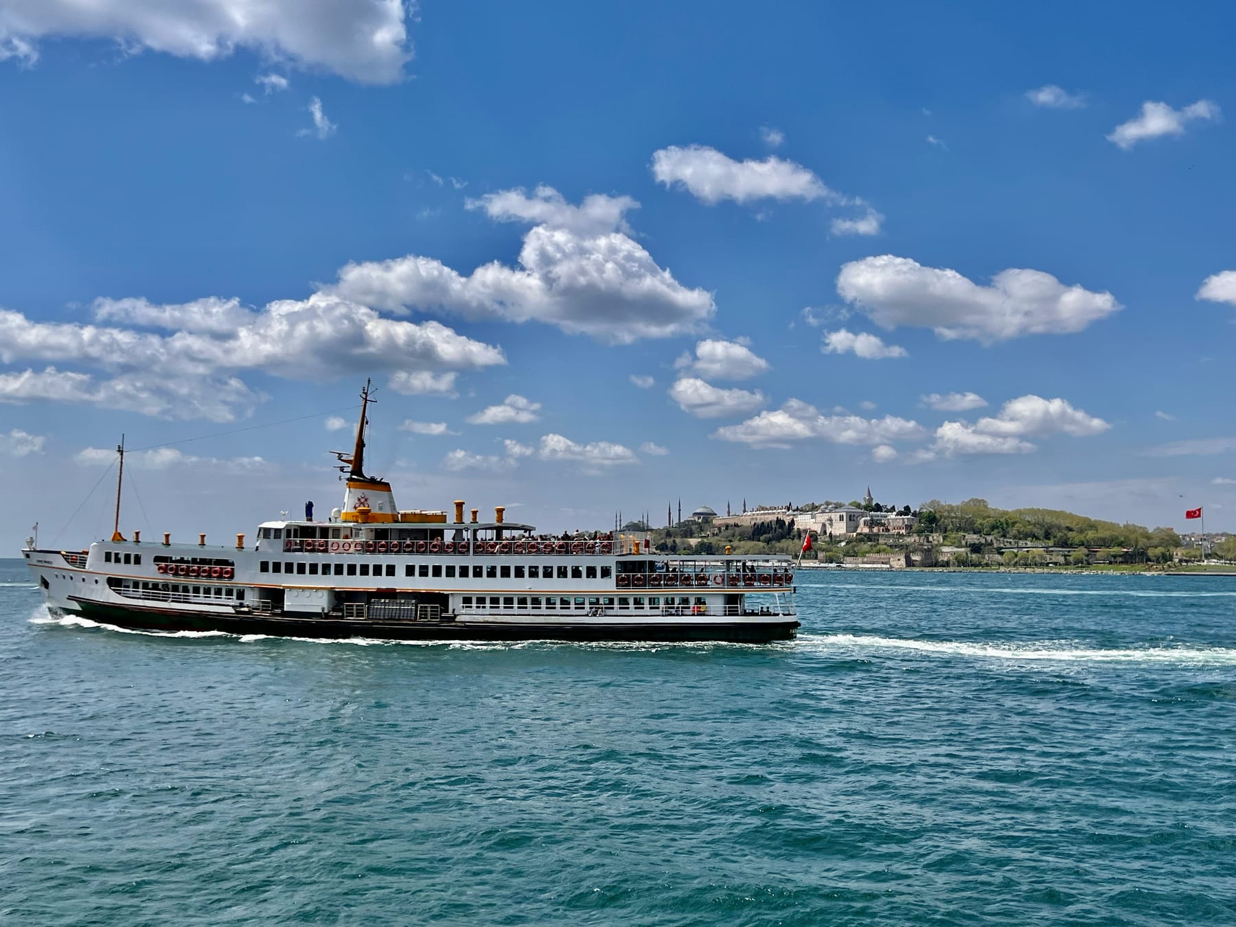 A classic Istanbul ferry crossing the Bosphorus on a sunny day, with Topkapi Palace and the Old City skyline on the shore behind